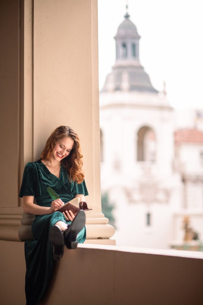 Author K.T. Jay sitting on a ledge writing with the view of Pasadena City Hall behind her.